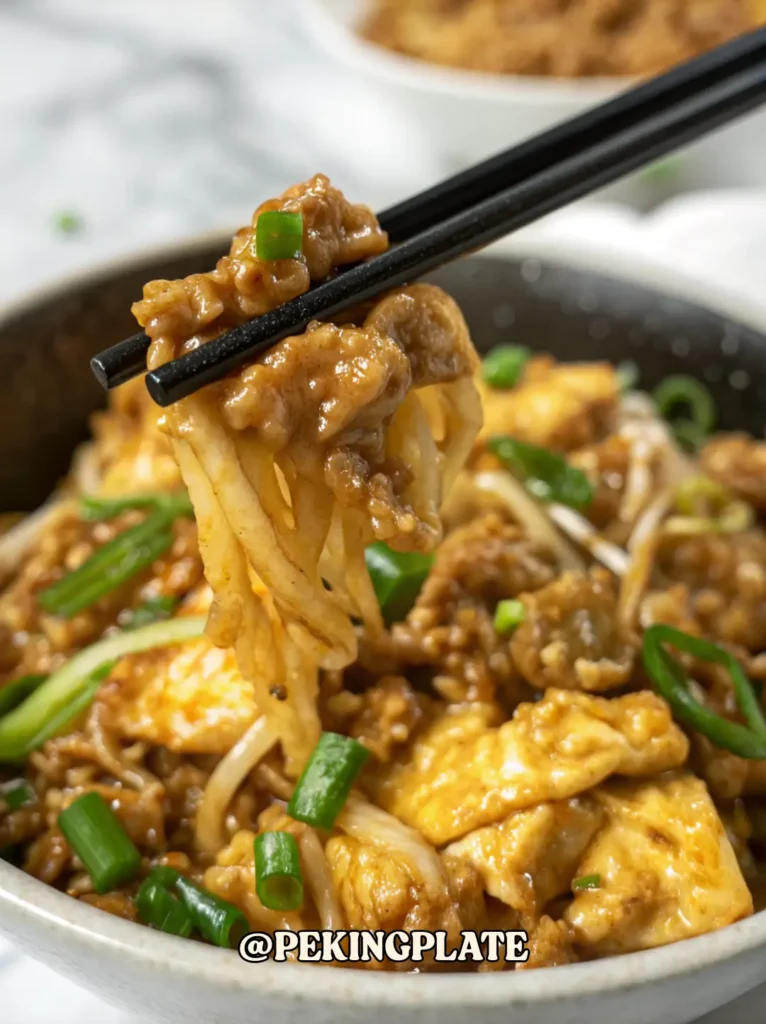 A close-up shot of black chopsticks lifting a bite of the easy stir-fried minced beef with enoki mushrooms and egg. The food is coated in a glossy sauce, showing the tender textures of the ingredients.